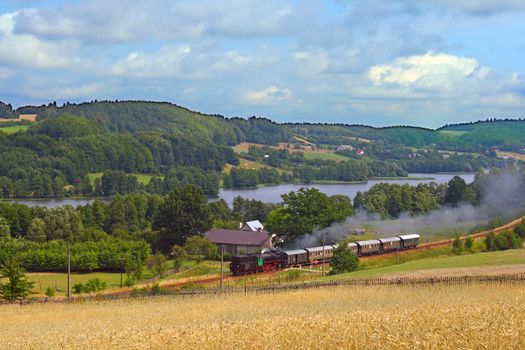 Rural summer landscape with retro train hauled by the steam locomotive running through the lakeside