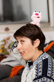 Young woman sticks out her tongue and has a doll on her head that sticks out her tongue too. 