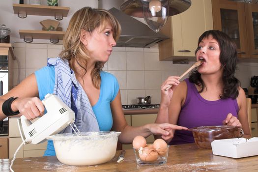 Pretty girls cooking together in the kitchen making cake and brownies. One girl is licking the chocolate of the spoon