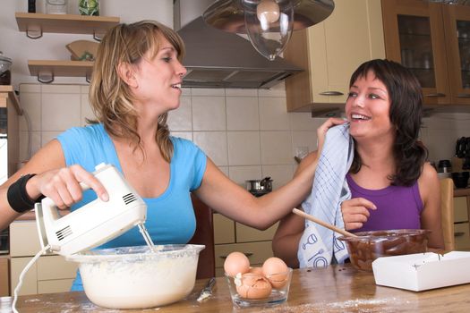 Two girls cooking together while one is having chocolate on her face. The other girl is wiping it away with a dishcloth