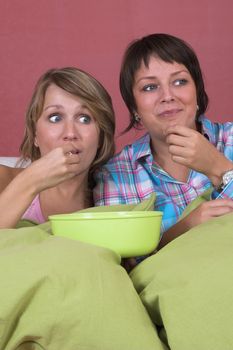 Two girls on the couch together watching a movie while eating popcorn and looking surprised