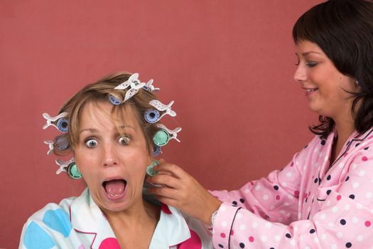 Pretty blond girl pulling a face of something being painful while her friend is putting hot rollers in her hair
