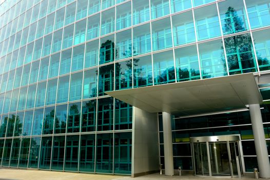 The deserted front door of a large commercial office block, surrounded by trees reflected in the glass. One tree reflection appears to grow out of a pillar of the entrance.
