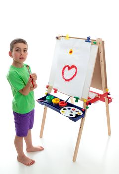 A young boy child stands by an easel with a beginning of a picture painted.  White background.