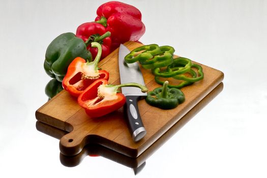 Red and green bell peppers being chopped at wooden cutting board with knife. Surrounded by hole bell peppers on reflective background