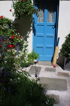 A cricket bat and ball resting against the wall and blue back door of a country cottage. Green foliage and a slate paving pathway surrounding.