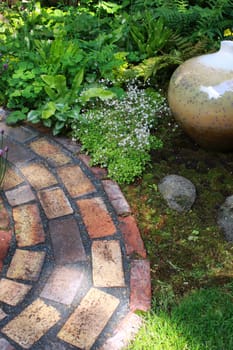 A section of an oval brick constructed garden path. Green grass, foliage and moss edge the path with an oval ceramic pot and stones.