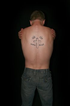 A topless man hugging himself, his back showing a crucifix tattoo.
