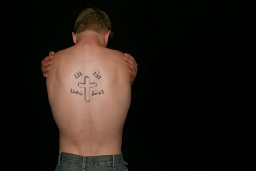 A topless man hugging himself, his back showing a crucifix tattoo.