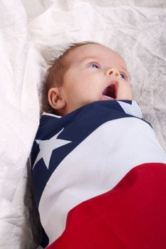 A cute baby wrapped up in a blanket with stars and stripes on it.  The baby is yawning.