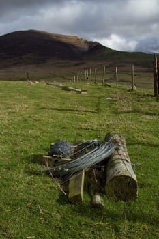 Agricultural landscape with wooden fence posts a coil of wire and a spool of barbed wire on green grass with a hill in the background.