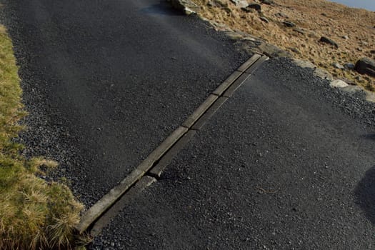 A tarmac footpath with stone curbs and grass with a sectioned drainage system spanning the width.