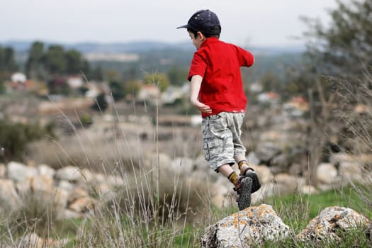 Little boy in red T-shirt and baseball cap jumps from the stone outdoors