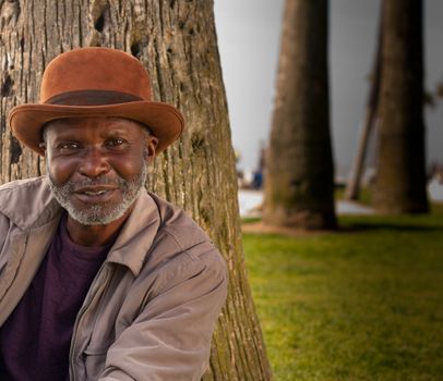 A happy man sitting on the beach.  There are trees in the background and some green grass.