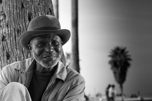 A photograph of a happy man relaxing against a tree at the beach.  There is people walking around in the background which adds to the excitement of the photogrpah.