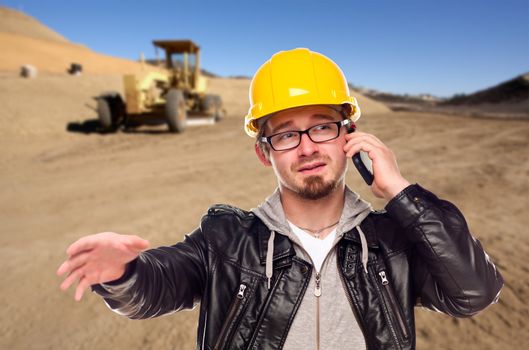 Young Cunstruction Worker on Cell Phone in Dirt Field with Tractor in the Background.