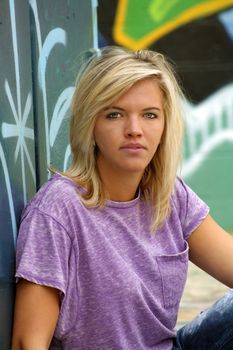 A lovely young blonde leans against a graffiti-covered garbage dumpster with a graffiti-covered wall behind her.