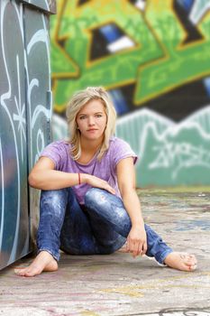 A lovely young blonde leans against a graffiti-covered garbage dumpster with a graffiti-covered wall behind her.