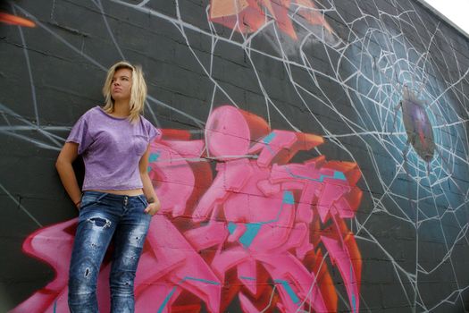 A lovely young blonde leans against a graffiti-covered wall.  Low angle.