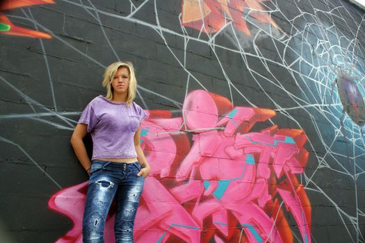 A lovely young blonde leans against a graffiti-covered wall.  Low angle.