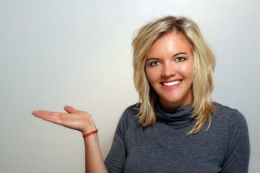 A studio close-up of a lovely young blonde with a bright, warm smile, holding her right hand out to frame left, palm up, pointing to your product or allowing you to place it in her hand.