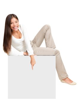 Woman sitting on blank empty paper poster, pointing down at copy space. Whole body image of cute casual woman in white isolated on white background.