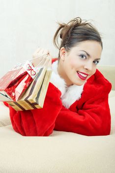 Smiling lady in the red furry Santa Claus costume laying indoors and holding bags with gifts