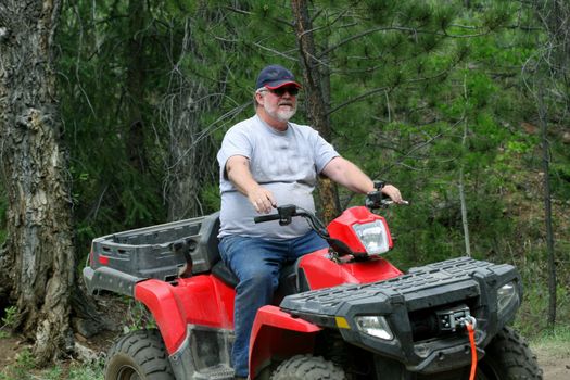 Bearded middle aged caucasian man with baseball cap riding red ATV in the forest.