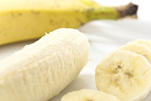 Close up shot of banana slices on a china plate, next to the remaining peeled banana portion, with an unpeeled banana in soft focus in the background. White background.