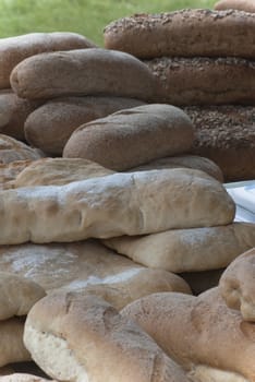 A selection of bread loaves and types on an outdoor market stall (not visible) with grass in soft focus in the background.  Mixture of white, brown and granary, seeded and unseeded.
