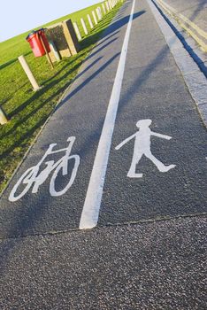 A British pavement, divided and signed for cyclists and pedestrians between an open field and a road.  Sunny day, blue sky.