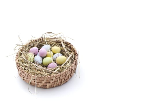 Coloured, speckled egg sweets laying on a bed of straw inside a circular basket, against a white background. Position:  lower left. Pastel egg colours:  pink, blue, green and yellow.