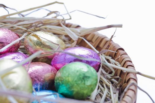 Egg shaped chocolates, covered in coloured foil, laying on a nest of straw inside a circular basket.  Pre-cropped.  Foil colours: gold, pink, purple, green and blue.  White background.