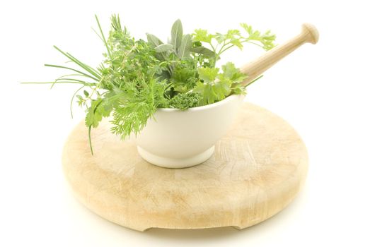 A selection of herbs inside a cream mortar with pestle, standing on light wood chopping board which shows signs of wear.  Isolated against white background.