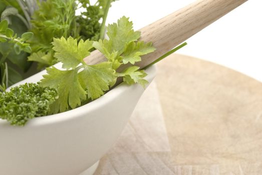 Closeup of a selection of herbs in a cream coloured mortar with pestle on worn wooden chopping board.  Subject cuts off at left edge of frame.  Isolated against white background.