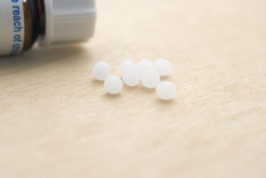 A closeup of homoeopathic pills resting on a light wooden surface with a closed pill bottle partly in-frame to the upper left (background).  Macro shot, shallow depth of field.