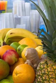 A selection of fruits laid out at a juice bar (stall), with cups and straws in soft focus in the background.  Includes bananas, oranges, apples, limes, lemons, ginger and pineapple.