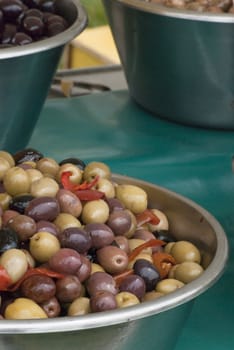 A bowl of dressed, mixed olives (green, purple and black) in a metal bowl on a market stall.  Others also in the background.  Rear of stall and grass visible. 