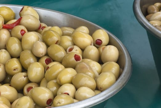 A metal bowl of green olives, stuffed with red chilli peppers, on a teal coloured tablecloth on a market stall.
