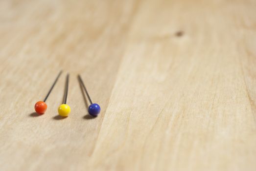 Close up of three steel pins with colourful plastic heads (orange, yellow and purple) lying on a birch wooden table in lower left frame, with empty table space to the right and above.  