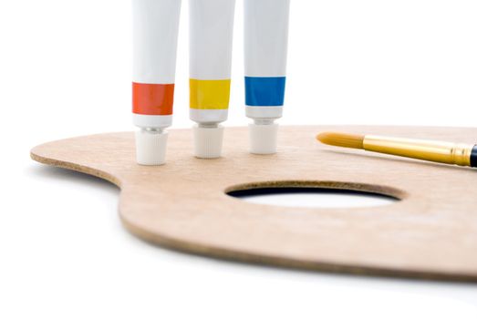 Three tubes of paint (primary colours) standing on edge of wooden palette with thumb hole and paintbrush tip visible.  Closeup shot, cropped in camera so only leftmost portion of palette is visible.  Isolated against white background.