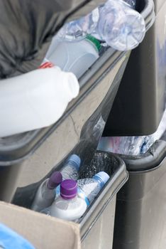 Overflowing black recycle bins, outdoors and awaiting collection, containing empty plastic bottles and packaging.  A box and a refuse sack are also visible.