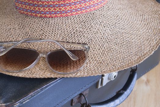 Close up of a 1980's style pair of sunglasses and a straw hat, resting on a battered, well-travelled small black suitcase.