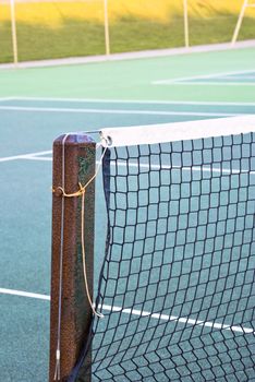 Tennis court net attached to rusting metal post with yellow and grey ropes.  Tennis court of green tarmac is visible.  Out of focus wire fence and shadowed grassy hill in background.  Winter daylight.