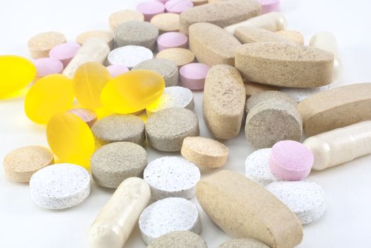 A mixture of different vitamin supplement pills, scattered on a white surface.  Includes hard tablets in various colours, also plastic and gel capsules.