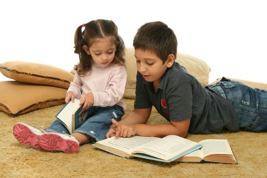 Brother and sister reading books over a carpet. They look interested and concentrated. 
