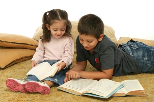 Brother and sister reading books over a carpet. They look interested and concentrated. 
