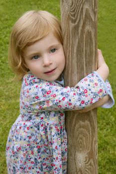 Beautiful little toddler blond girl hug a trunk with grass park background