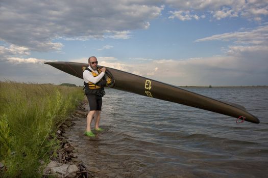 mature paddler is carrying  a long racing carbon fiber kayak to launch it on a lake with grassy and rocky shore in northern Colorado (thirteen - temporary race number placed on deck by myself)