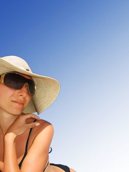Woman in bikini, hat and sunglasses lying on beach over gradient blue sky